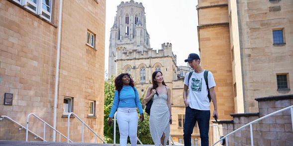 A group of three students on campus. The students are walking in between two stone buildings with a historic tower in the background. 